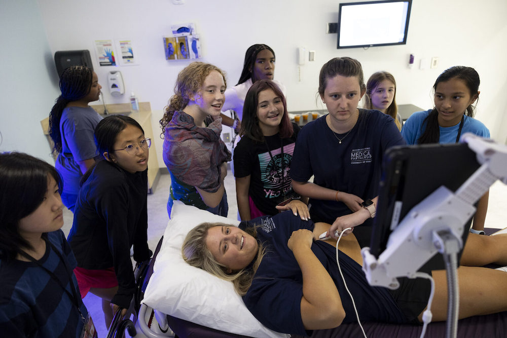 (L-R) Medical Partnership M2 student Jordan Beil has an ultrasound used on her by fellow M2 student Amanda Lilley as local students watch the demonstration during the Mini-Med Camp at the Medical Partnership.